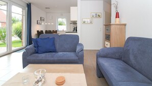 Ein modernes Wohnzimmer mit zwei grauen Sofas, einem Holztisch und einem Glasbehälter mit Muscheln. Im Hintergrund ist eine Essecke mit einer Küche erkennbar.