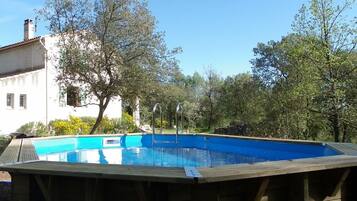 Ein hölzerner Pool mit blauer Innenseite, umgeben von Bäumen und einem weißen Gebäude im Hintergrund.