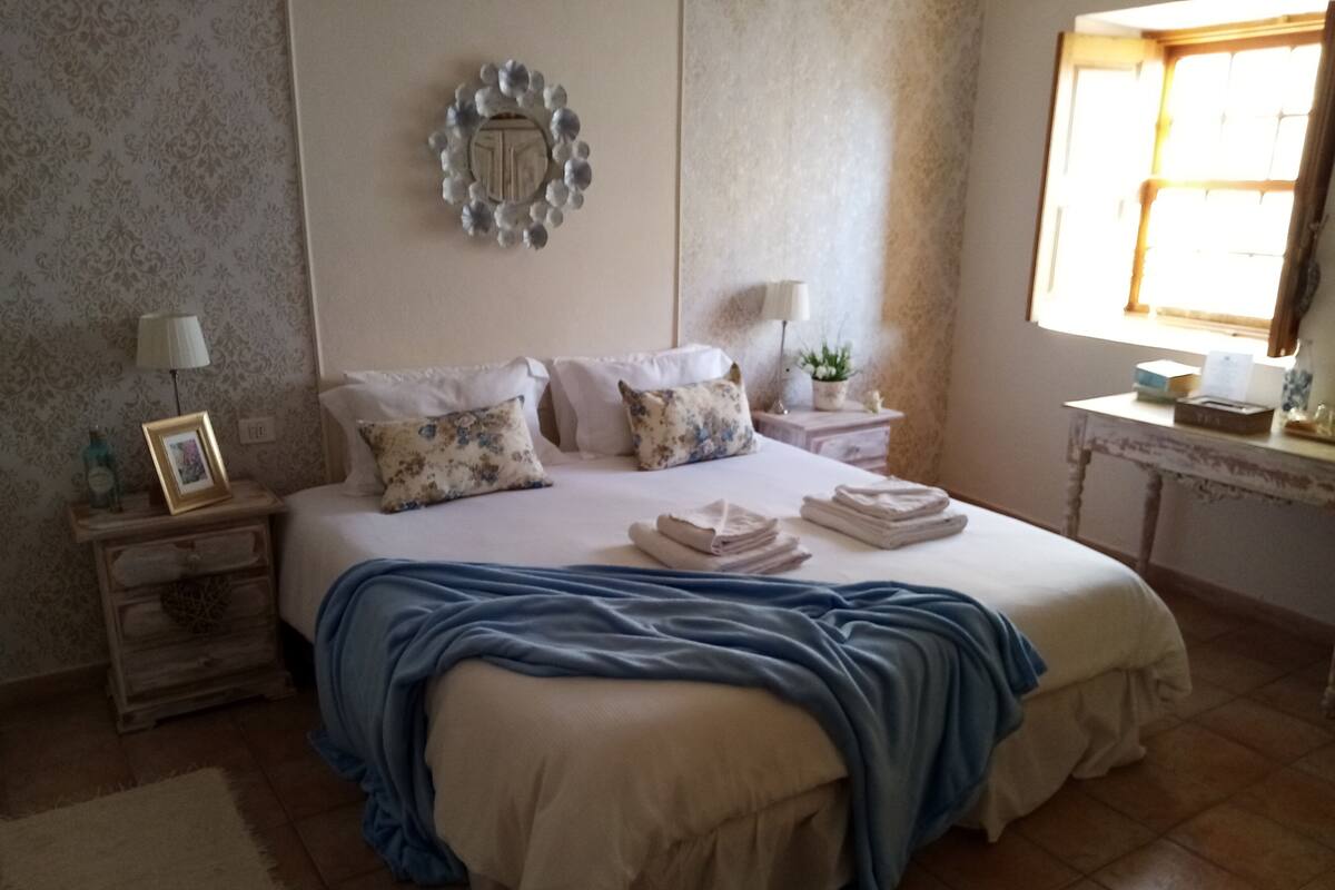 Ein Schlafzimmer mit einem Bett, Kissen, einem Nachttisch, einer Lampe, einem Fenster und einer Wanddekoration.