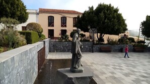 A statue in a courtyard with a fountain, benches, and a building with arched windows in the background.