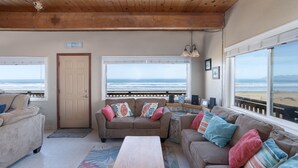 A cozy living room with a wooden ceiling, a beige couch with colorful pillows, a wooden coffee table, and large windows overlooking the beach. - Steps from the Sand! Spacious Beach Home with Amazing Ocean Views! (Oceano)