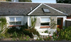 Ein Haus mit geneigtem Dach, einem Dachfenster und einer kleinen Terrasse mit weißen Stühlen.