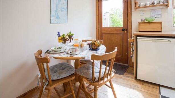Un comedor con una mesa de madera dispuesta para cuatro personas, rodeada de sillas con cojines. Se ve una puerta de madera y un refrigerador al fondo.