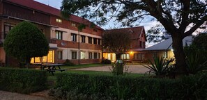 A multi-story building with a brick facade, a covered patio area, and a well-maintained garden with a picnic table. - Lovasoa CCCC - Hostel (Antsirabe)