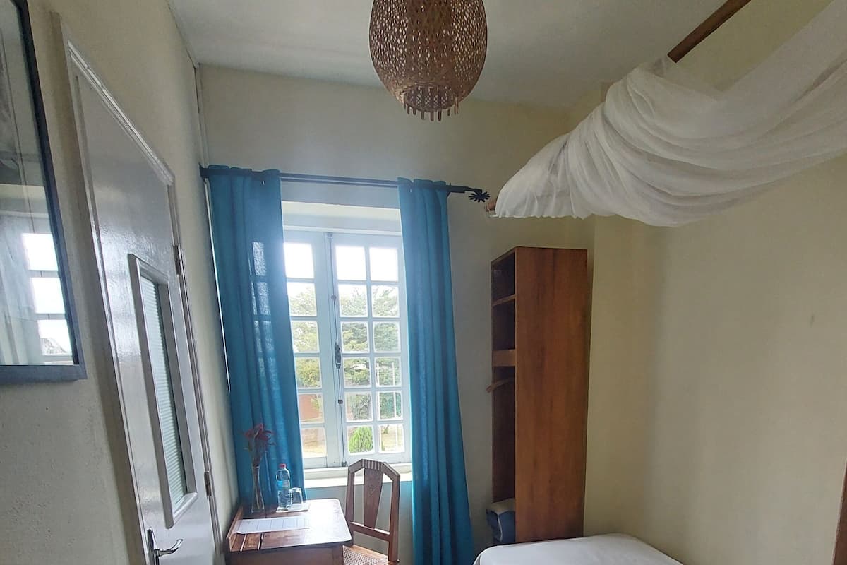 Ein kleiner, sauberer Raum mit einem Bett, einem Schreibtisch, einem Stuhl, einem Fenster mit Vorhängen und einer Pendelleuchte.