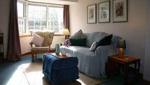 Un salon chaleureux avec un canapé, un fauteuil et un pouf, une fenêtre avec des rideaux et des œuvres d’art encadrées accrochées au mur.