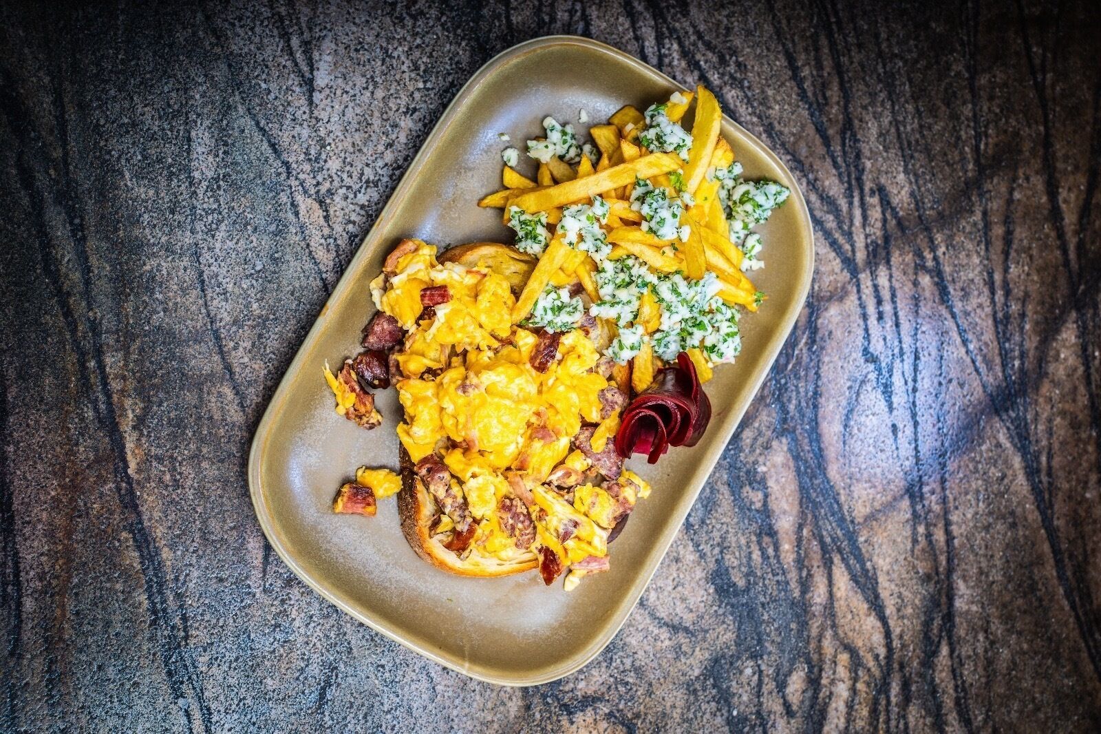 A rectangular plate with scrambled eggs, bacon, French fries, and a side salad on a textured surface.
