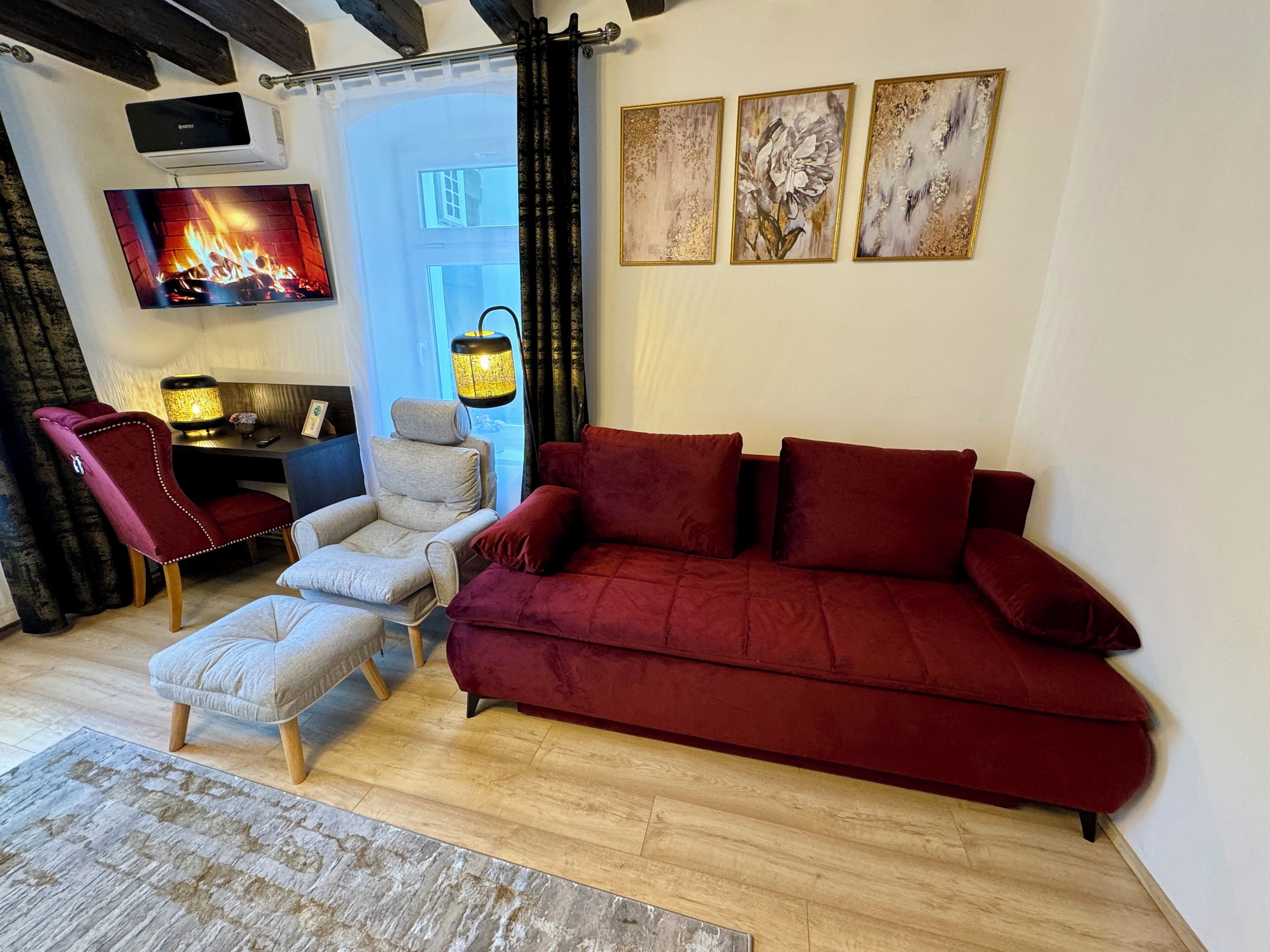 A cozy living room with a red sofa, a grey armchair, a small ottoman, a TV displaying a fireplace, and three framed artworks on the wall.
