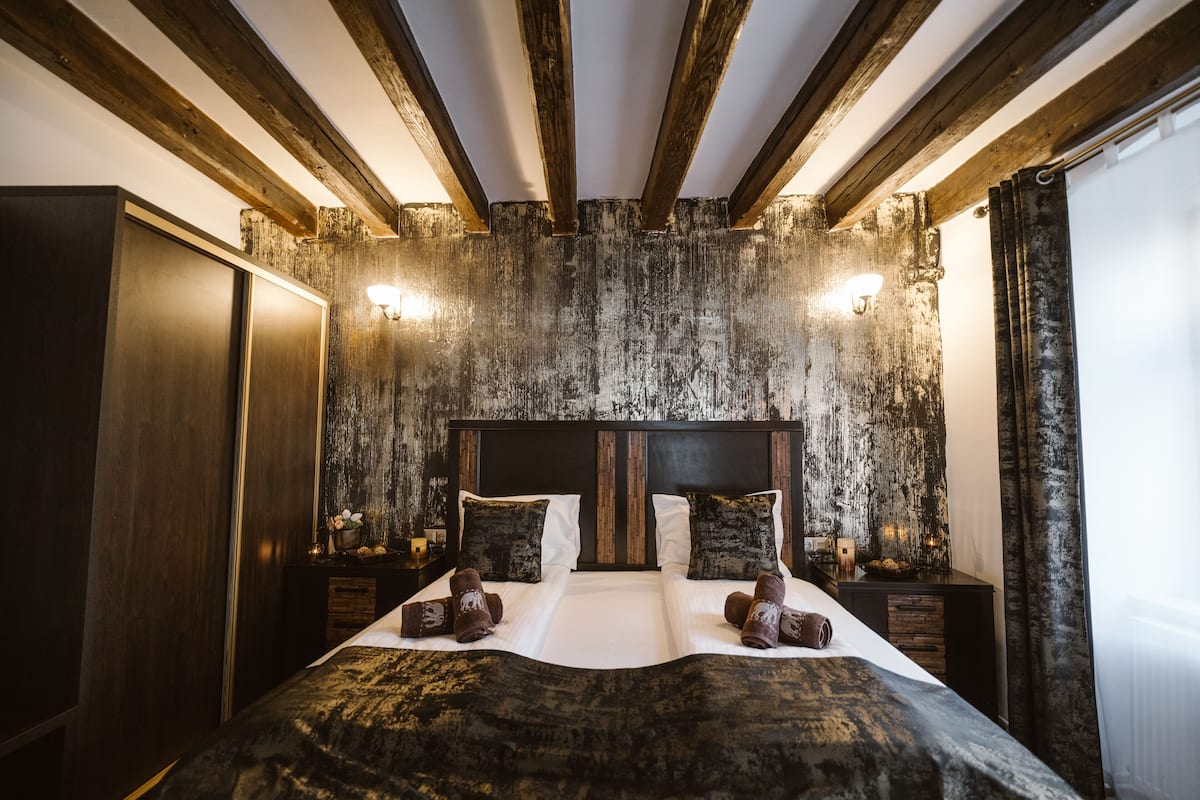 A large bed with a dark, textured bedspread and two pillows, wooden beams on the ceiling, and wall-mounted lights.