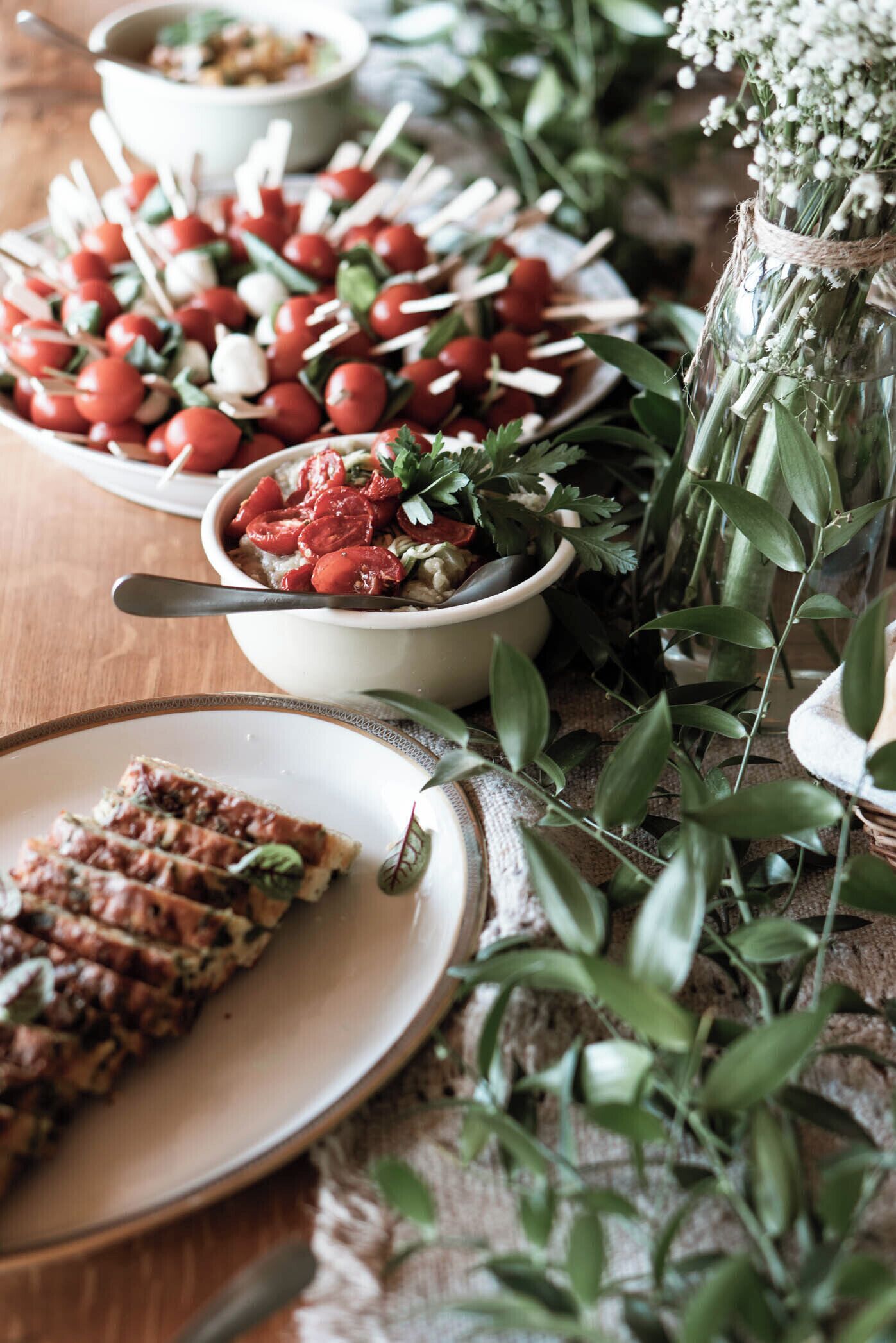 Una mesa con varios platos, incluyendo un tazón de tomates cherry y vegetales, un plato de carne en rodajas y una jarra de cristal con flores.