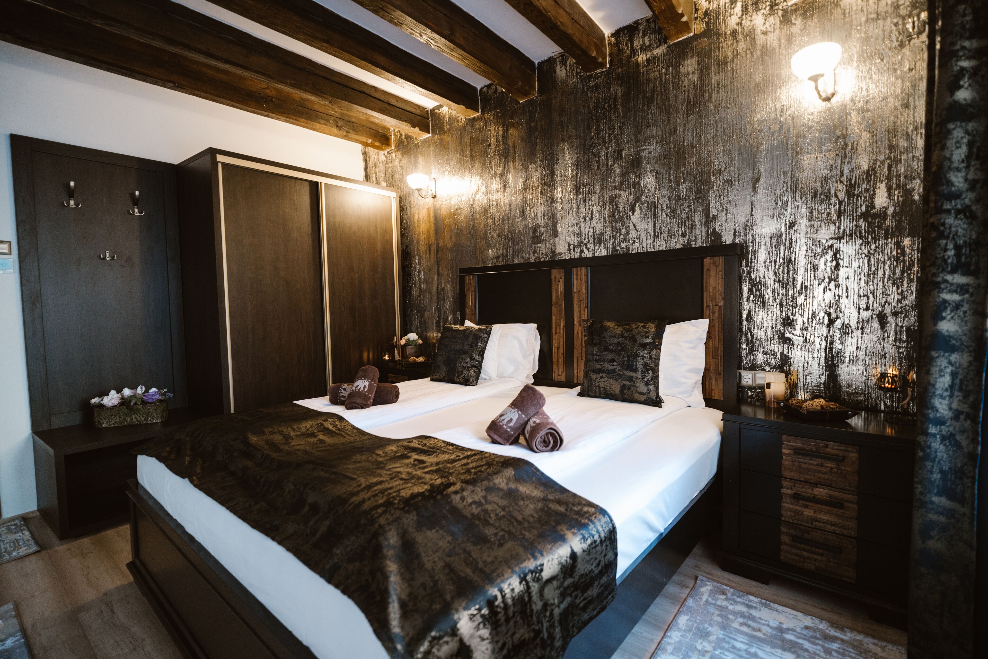A hotel room with a large bed, wooden headboard, and a rustic wall. There are two bedside tables with lamps and a wardrobe.