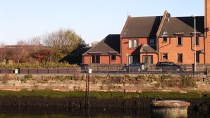 A residential building with a brick facade, a car parked in front, and a body of water in the foreground. - Sunny south facing Riverside home  (Ayr)