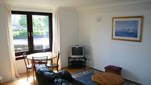 A living room with a black leather sofa, a wooden coffee table, a TV on a stand, a dining table with chairs, and a window with curtains. - Sunny south facing Riverside home  (Ayr)