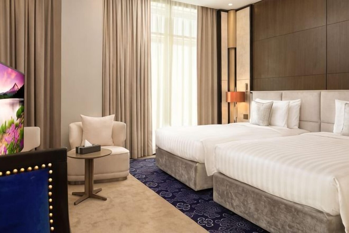 Habitación de hotel con dos camas, una mesita, una silla, un televisor y un ventanal con cortinas.