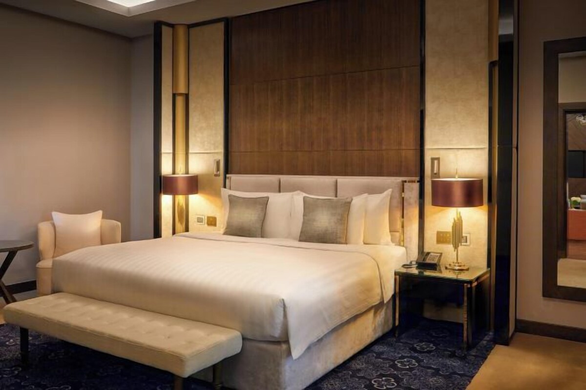 Amplia habitación de hotel con una cama grande, un banco, dos mesitas de noche con lámparas y un cabecero de madera.