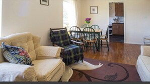 Una sala de estar con un sofá, un sillón escocés, una mesa de comedor con sillas y una zona de cocina al fondo.
