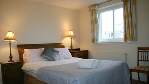 A neatly made bed with a white comforter and pillows, two bedside lamps, a wooden nightstand, a window with yellow curtains, and a wooden chair.