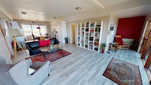 A spacious living room with a black leather sofa, a grey armchair, and a red rug. There is a bookshelf filled with books and a dining area with a table and chairs. - Kleifar (Hellissandur)