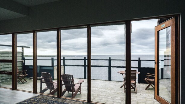 A room with large glass doors leading to a balcony with a view of the sea. There are several wooden chairs and a small table on the balcony. - Kleifar (Hellissandur)