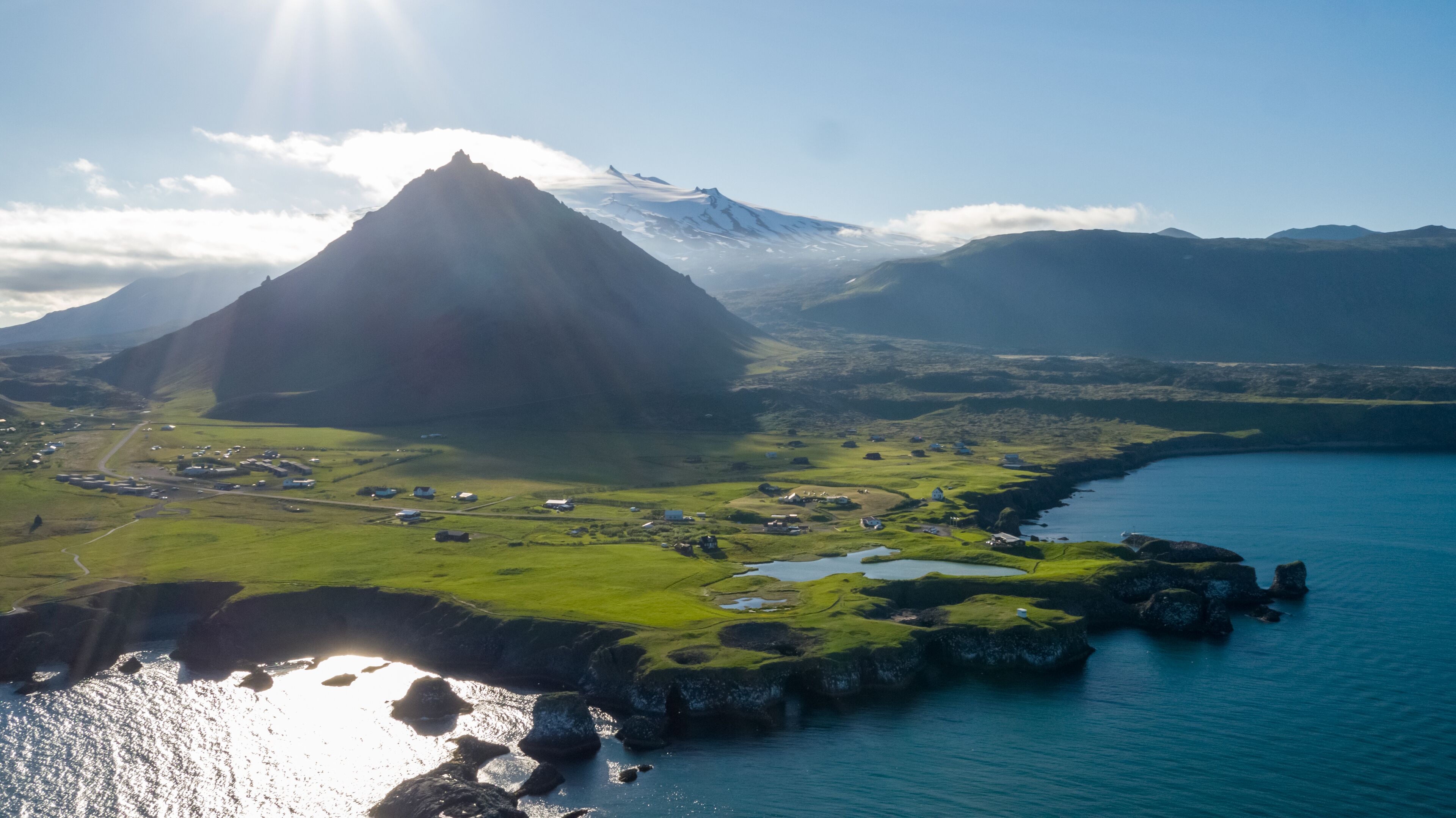 Paisagem costeira com uma montanha imponente, campos verdes e um pequeno povoado à beira-mar.