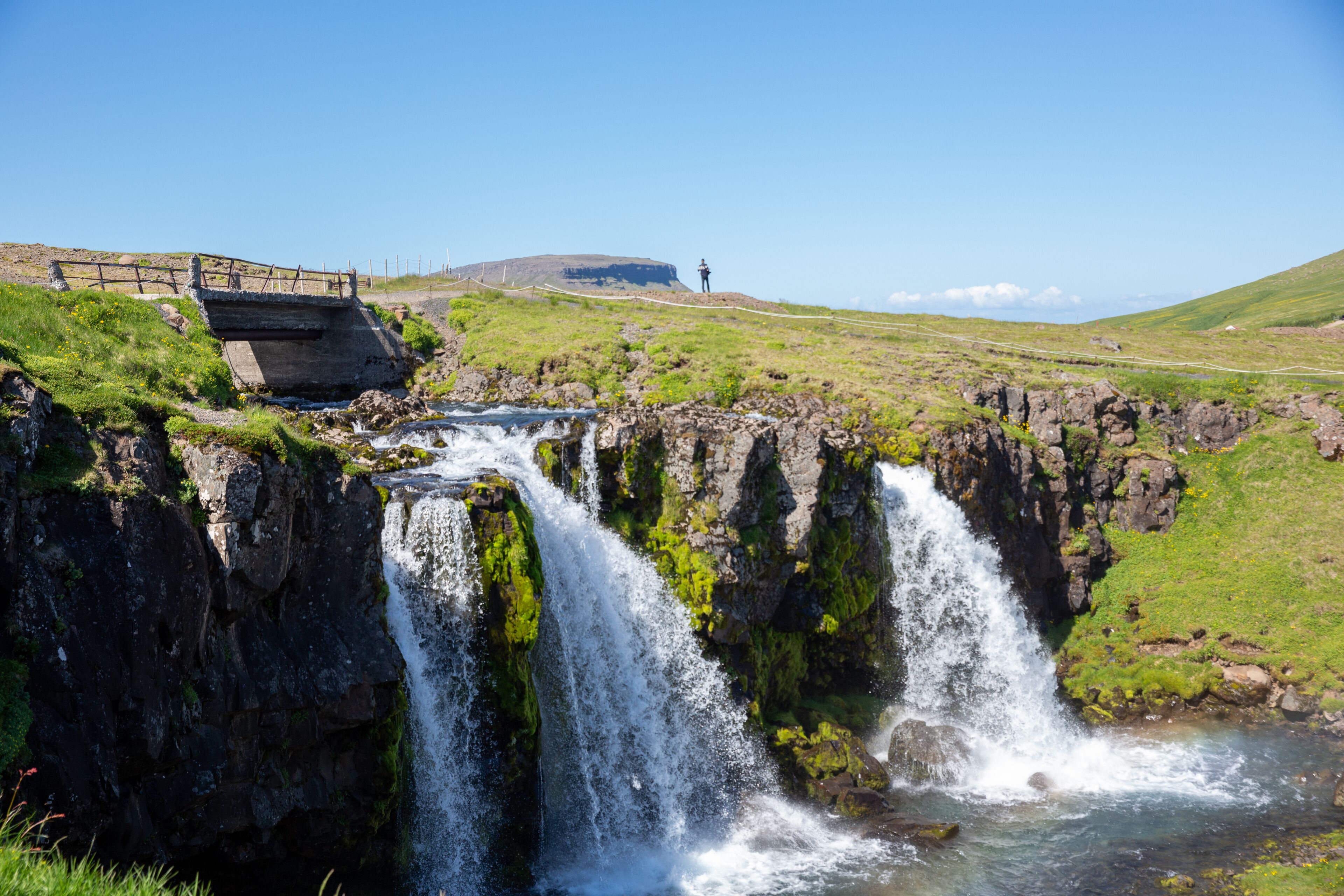 A waterfall cascading over rocky terrain with a bridge and a distant hill.