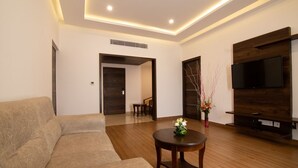 A modern living room with a sofa, a small round table, a TV mounted on a wooden wall, and a door leading to another room. - Liverpool Hotels, Outer Ring Road, Marathahalli (Bengaluru)