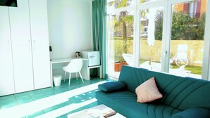 A modern living room with a green sofa, a white coffee table, and a small white desk with a chair. There is a large sliding glass door leading to an outdoor area with a garden and a bicycle. - Villa Marina (Venice)