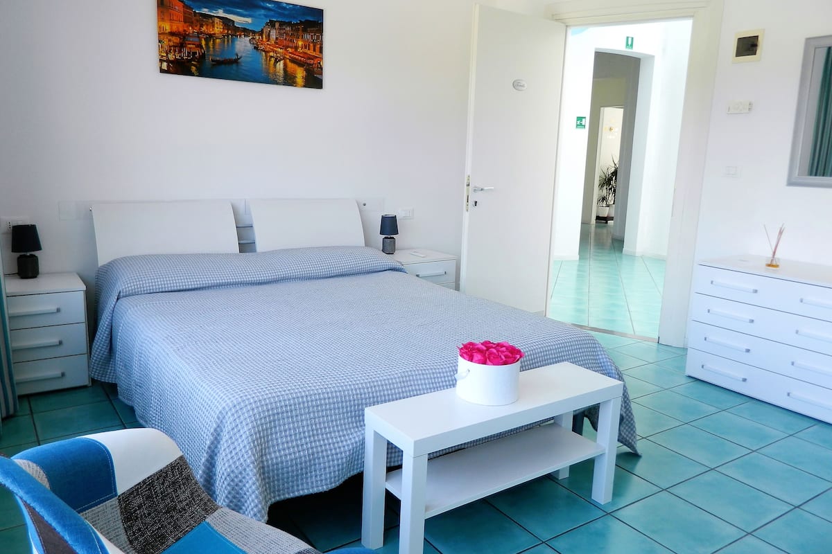 A hotel room with a bed, a bedside table with a vase of flowers, a chair, a dresser, and a painting on the wall.