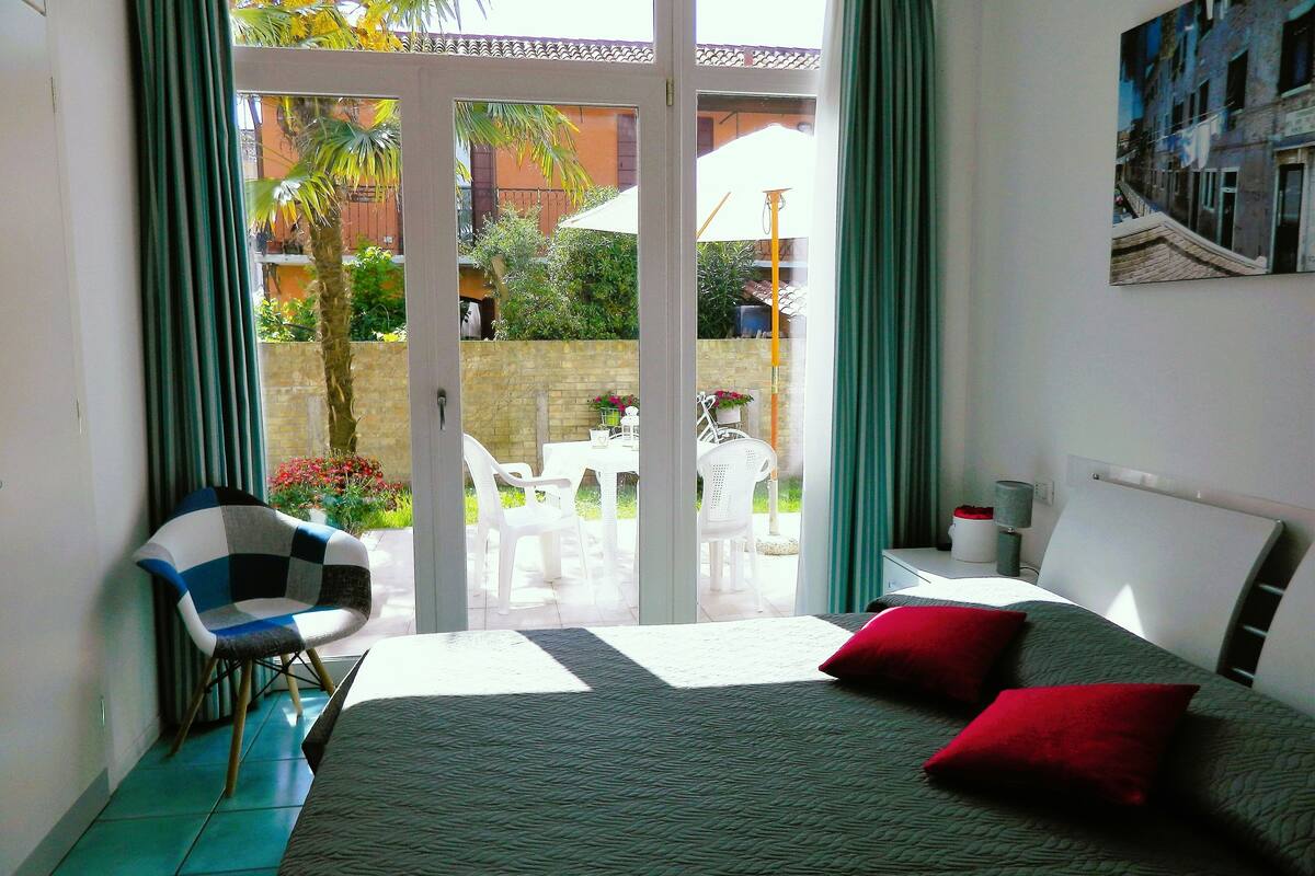 A bedroom with a bed, two red pillows, a white chair, and a table with chairs outside a sliding glass door.