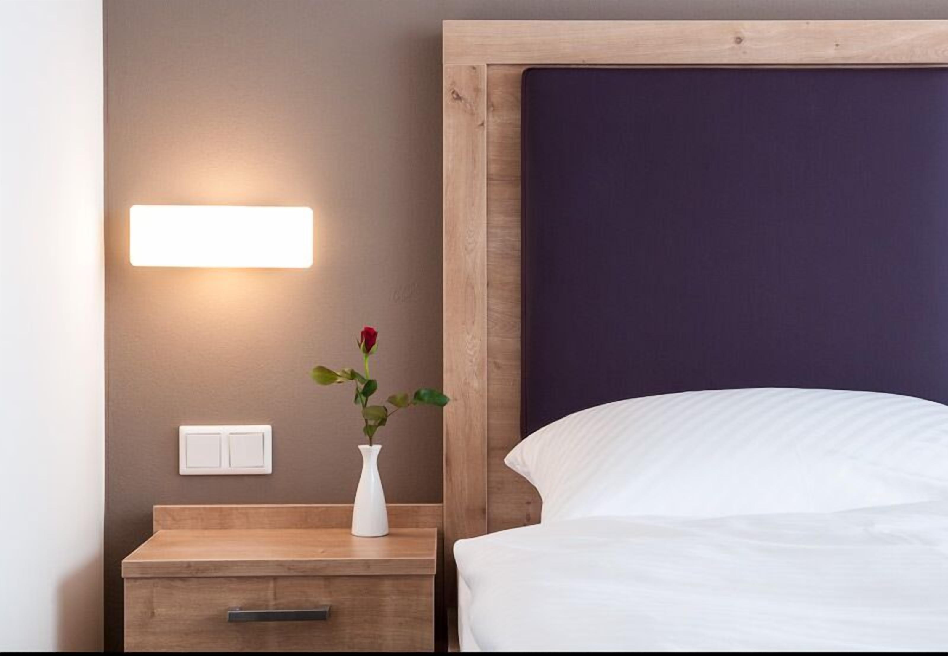 Ein ordentlich eingerichtetes Hotelzimmer mit Nachttisch aus Holz, einem Beistelltisch, einer weißen Vase mit einer roten Rose, einem Bett mit weißer Bettwäsche und einer Wandleuchte.
