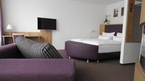 A modern hotel room with a bed, a sofa, a TV, and a small table with a lamp.