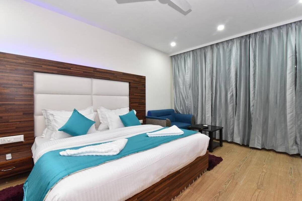 Ein modernes Hotelzimmer mit einem großen Bett, einem Holz-Kopfteil, einem blauen Sofa, einem kleinen Tisch und grauen Vorhängen.