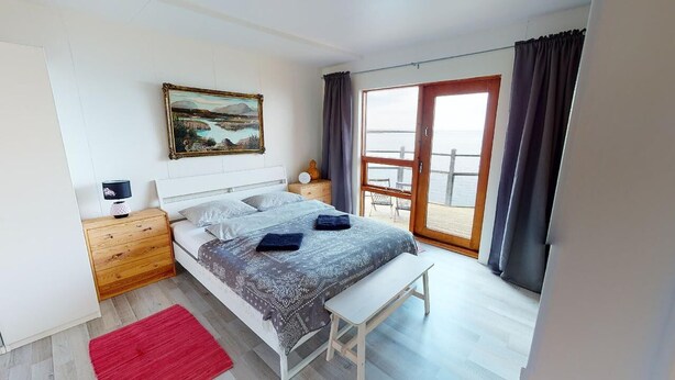A bedroom with a bed, bedside tables, a bench, a painting, and a view through a large window.