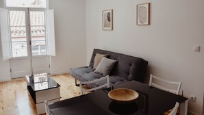 A modern living room with a sofa, a dining table, and a window with a view of buildings. - Spacious, light-filled one-bedroom apartment (self check-in) (Coimbra)