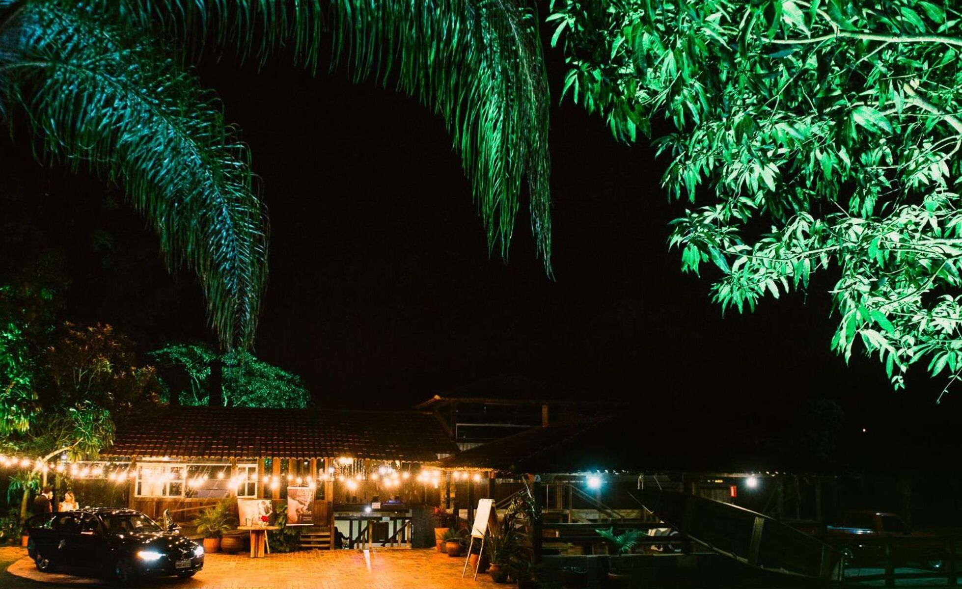 Vista noturna de um edifício rústico com luzes de corda, um carro e vegetação ao redor.