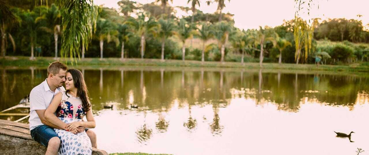 A couple sitting by a calm lake with a wooden dock, surrounded by lush greenery and palm trees.