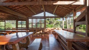 Un espacio de comedor techado con bancos y mesas de madera, ventanas amplias y vista a zonas verdes.