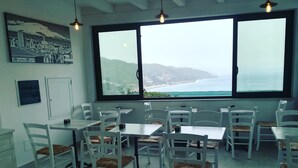 Un espace repas avec des tables et des chaises blanches, de grandes fenêtres offrant une vue sur la mer et les collines, et des luminaires suspendus.