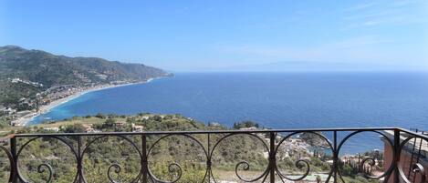 Varanda com cadeiras de metal e uma pequena mesa, com vista para a paisagem costeira e uma visão nítida do mar e edifícios ao longe.