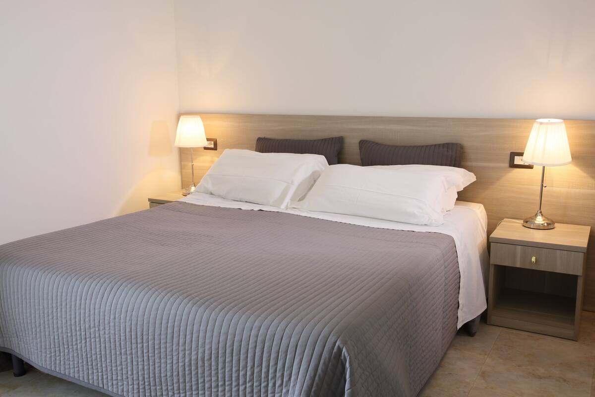 Una cama bien hecha con colcha gris, dos mesitas de noche con lámparas y un cabecero de madera.