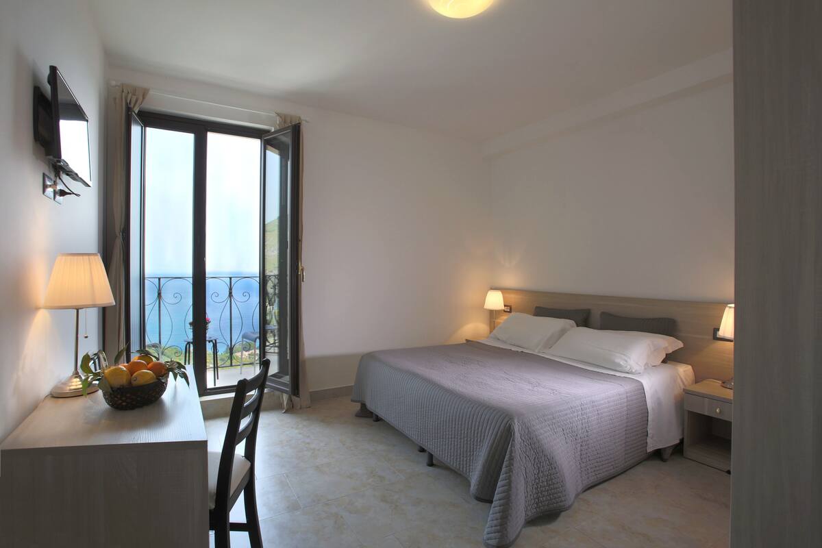 Habitación de hotel con cama, mesitas de noche, un escritorio con lámpara, frutera y vistas al mar a través de un ventanal grande.