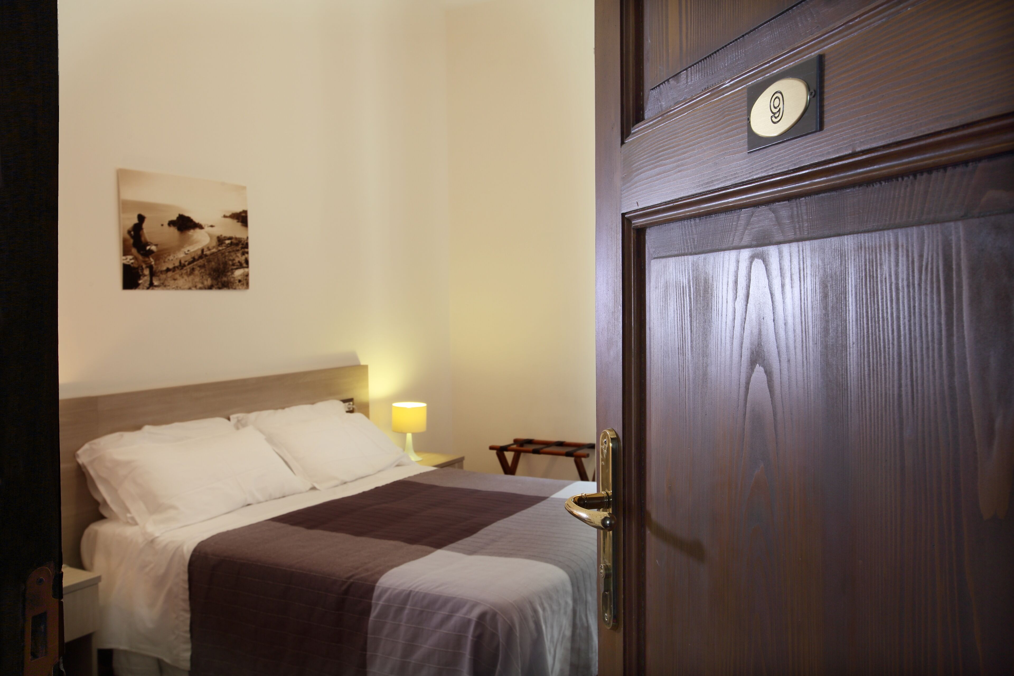 A hotel room with a bed, a nightstand with a lamp, and a door with a number plate.