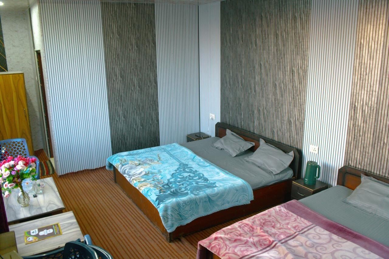Camera d'albergo con un letto singolo, un comodino con una tazza verde, un tavolino di legno, un piccolo tavolo con un vaso di fiori e una sedia.