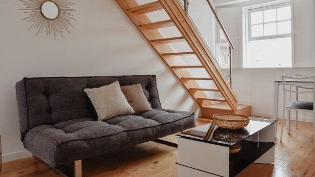 Een moderne woonkamer met een grijze bank, een houten trap en een groot raam dat voor natuurlijk licht zorgt.