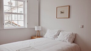 Een net opgemaakt bed met witte lakens, een bedlampje en een ingelijste foto aan de muur.