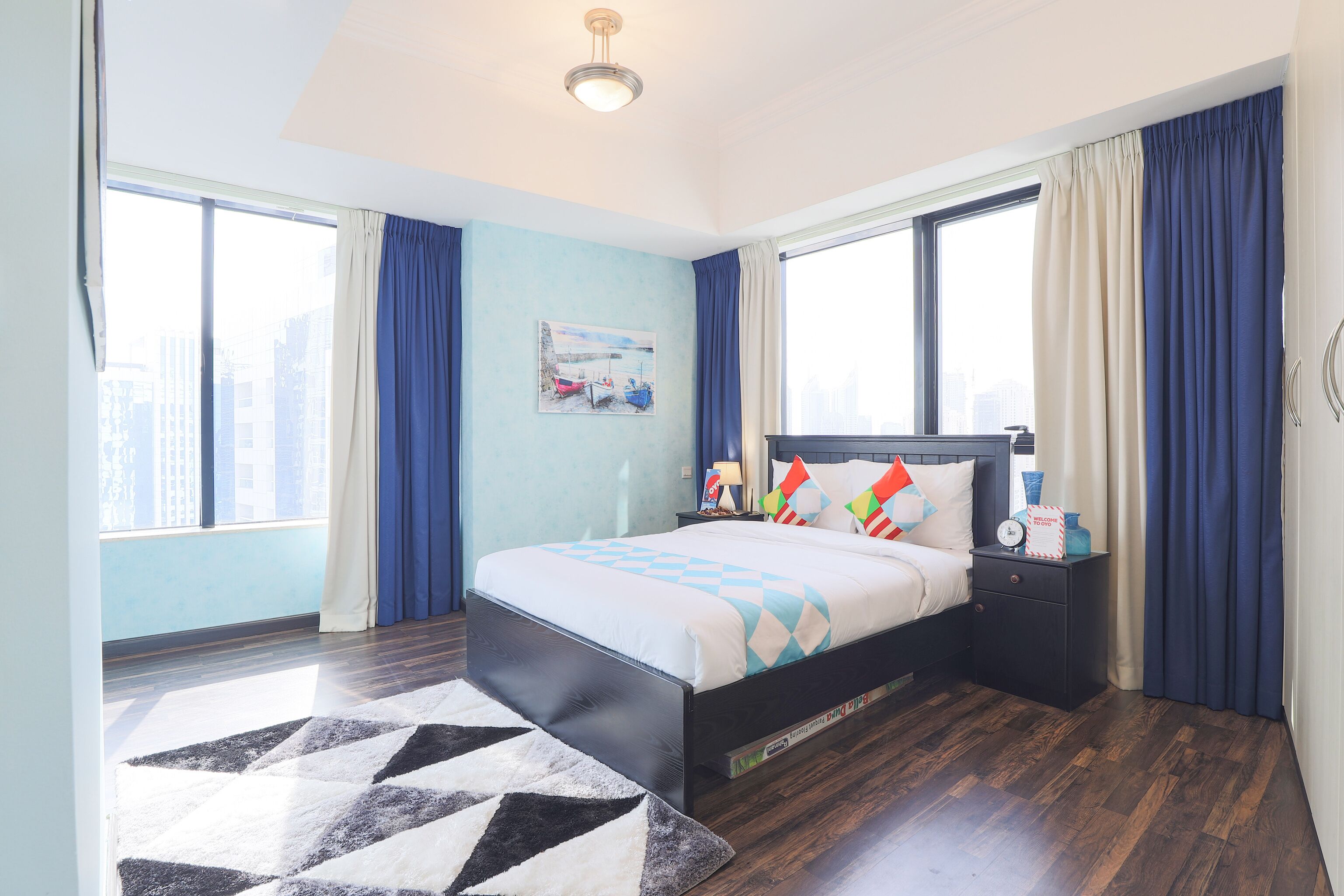 A modern bedroom with a large bed, a nightstand, a painting on the wall, and a view of the city through the windows.