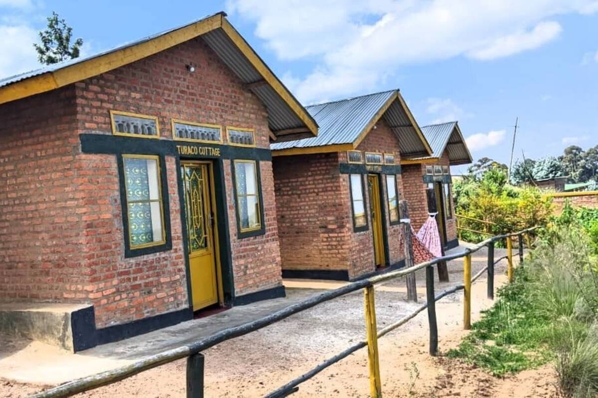 A brick building labeled "Turaco Cottage" with a yellow door and windows, a metal roof, and a wooden fence in front.