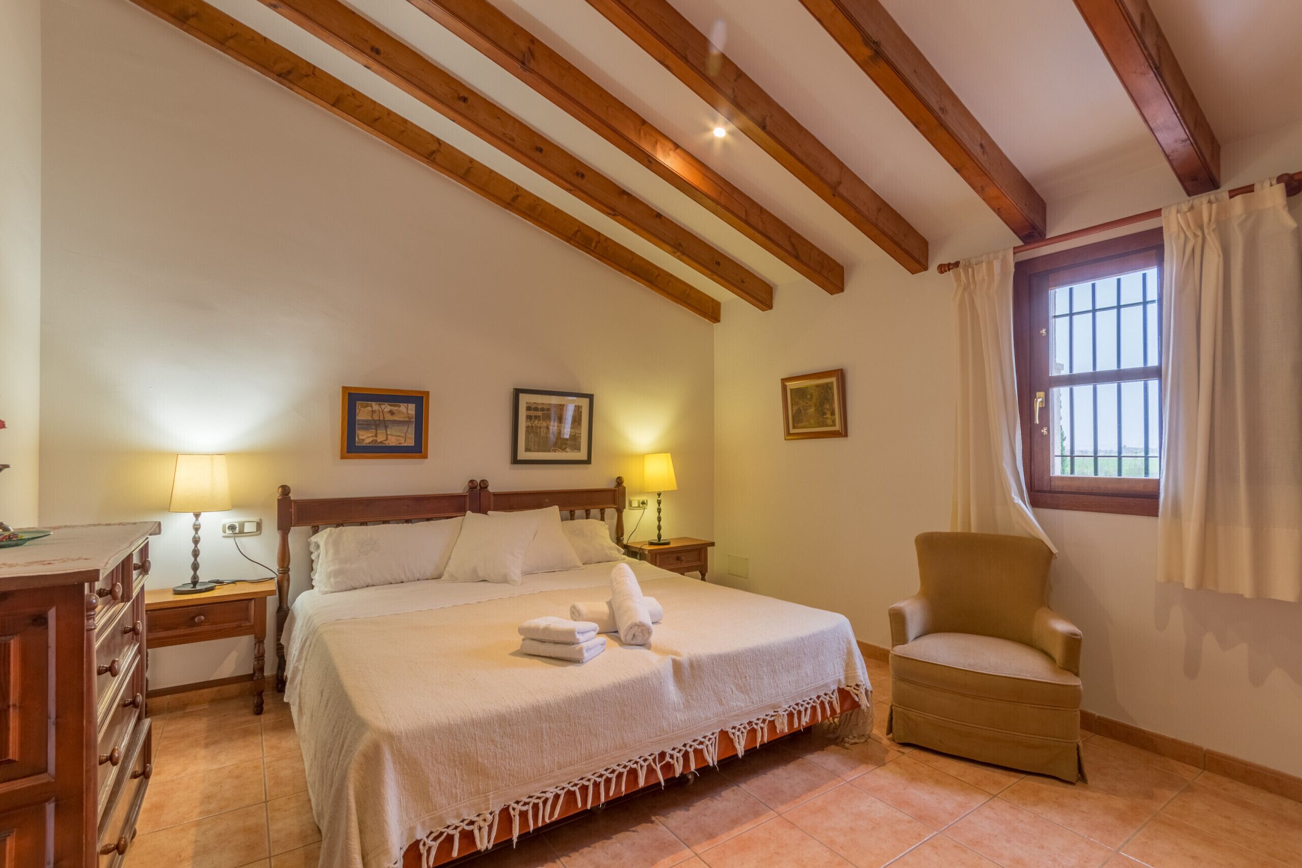 Une chambre d’hôtel avec un plafond en bois, un lit recouvert de draps blancs, deux lampes de chevet, une chaise et une fenêtre avec des rideaux.