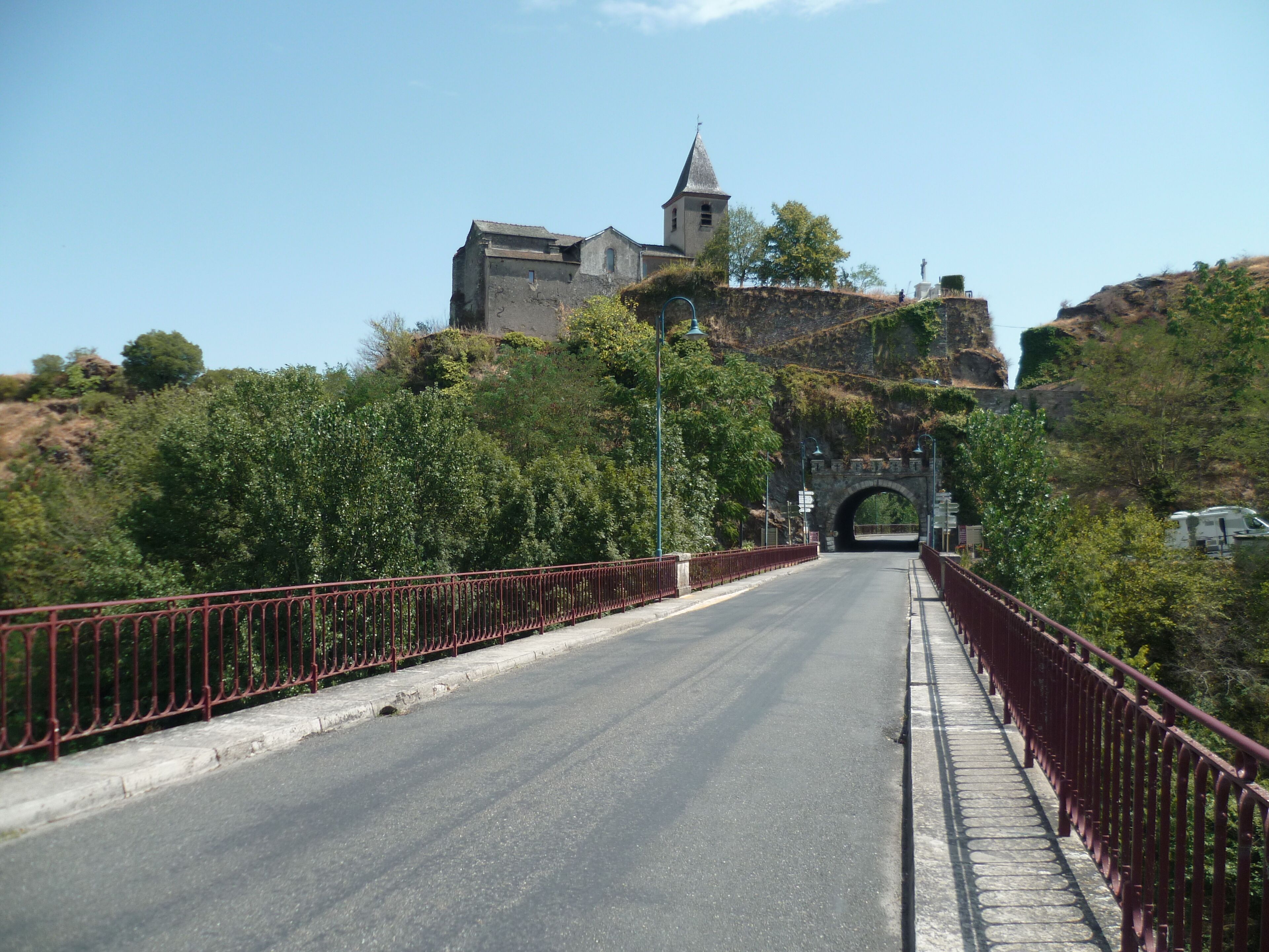 A road leading to a historic building with a tower, surrounded by greenery and a stone bridge.