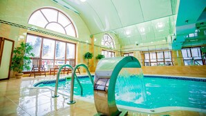 Una piscina interior con paredes de vidrio transparente, suelo de baldosas y una zona de estar con sillas y mesas.